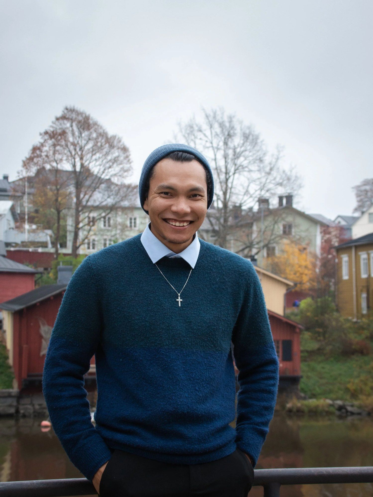 Hymyilevä mies pipossa ja neuleessa punaisten puutalojen edessä